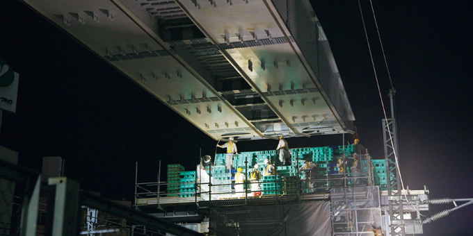 橋脚の上では作業員が緑色の高強度ブロックを積み上げ，伸びてくる架設機の走行ガーダー先端を受けるの写真