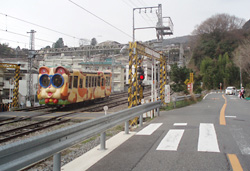 現在の同じ場所。踏み切りの道は狭く、車一台通れる程度だが、線路の向うには住宅街が広がる。山の形と山に向かって伸びる線路は変わらない。山頂の遊園地に合わせたデザインの電車となっている。これは猫の「ミケ」