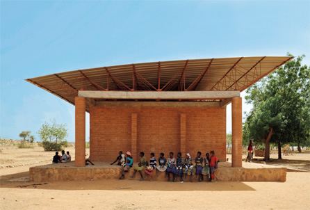 写真：鋼板の屋根とレンガづくりの教室との間に，トラス構造の空隙が見える。これによって熱気を外に逃がしている。大きな屋根は教室の周囲にも日陰をつくり出す