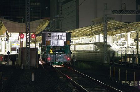 4両編成が動力台車を先頭に南部ヤードから出発。東京駅ホームに留め置かれた新幹線車両の隣りを通り抜けるの写真