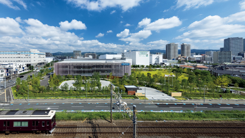 図版：阪急京都線沿いに正対した南東の構え。芝生広場や緑溢れるウォーキングコースなど公園のように地域にひらかれた「コミュニケーションパーク」