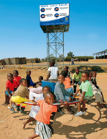 写真：ポンプで遊ぶ子どもたち。ポンプを回すと背後上空のタンクへと水が吸い上げられる。タンクを囲う看板には保健省の広告が掲げられている