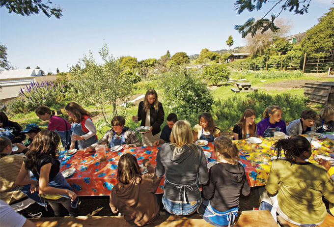 写真：キッチンの前の木陰に長さ20mのダイニングテーブルがあり，クラス全員がテーブルを囲んで食事を楽しむ 