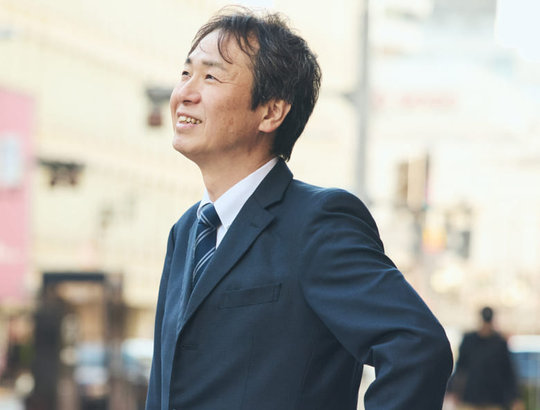 明るい屋外で紺色のスーツをまとい、晴れやかな表情で空を見上げる右上肢障がいの男性社員の写真