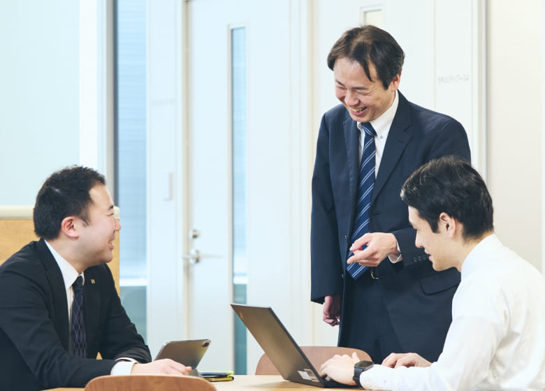 会議室で資料を囲んで打ち合わせをする右上肢障がいの男性社員と同僚たち。身振り手振りを交えてフランクに会話する業務風景の写真