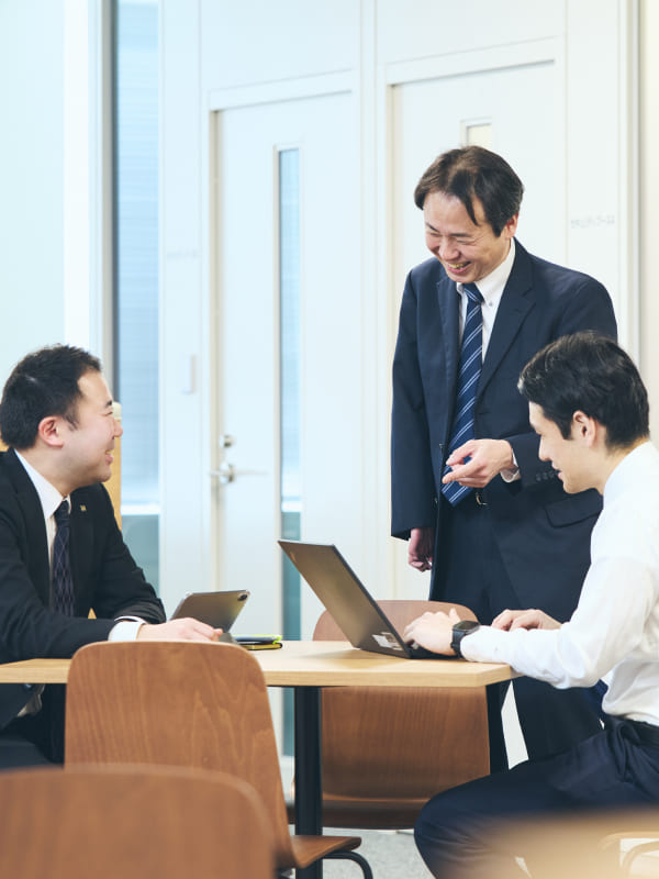 会議室で資料を囲んで打ち合わせをする右上肢障がいの男性社員と同僚たち。身振り手振りを交えてフランクに会話する業務風景の写真