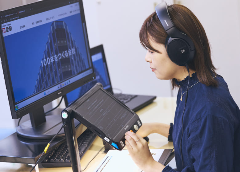 ヘッドホンを装着し、専用の拡大読書器を指先で操作しながら、集中してPC業務に取り組む視覚障がいの女性社員の写真