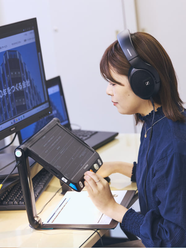 ヘッドホンを装着し、専用の拡大読書器を指先で操作しながら、集中してPC業務に取り組む視覚障がいの女性社員の写真