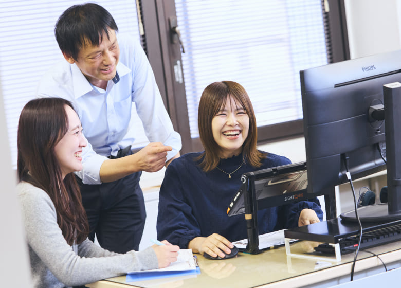 モニターを前に同僚と談笑し、はじけるような笑顔を見せる視覚障がいの女性社員の写真