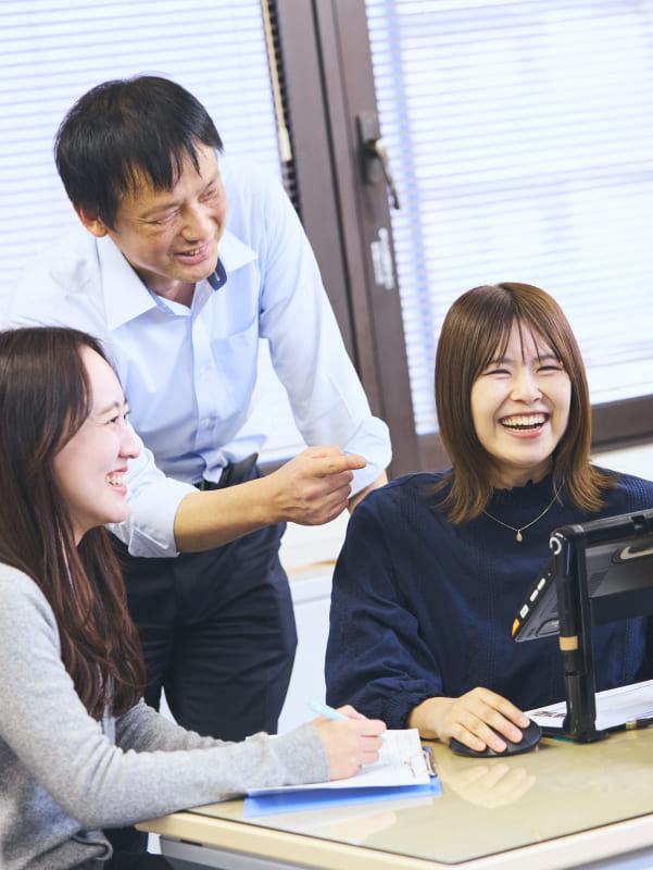 モニターを前に同僚と談笑し、はじけるような笑顔を見せる視覚障がいの女性社員の写真